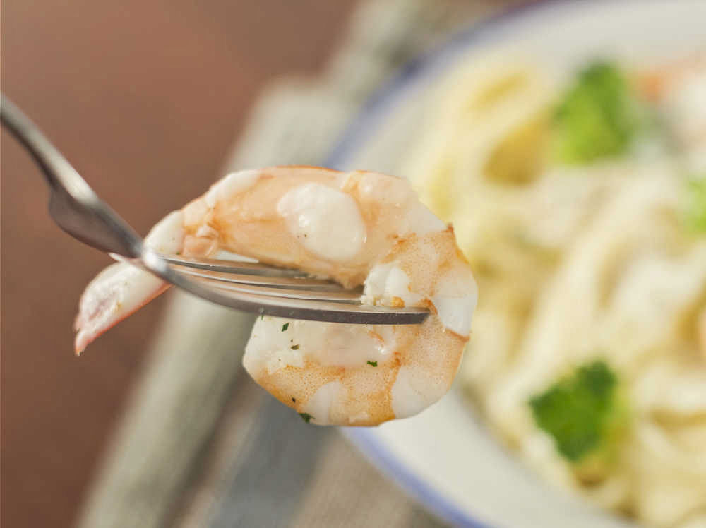 Shrimp on a fork in front of a shrimp pasta with broccoli dish.
