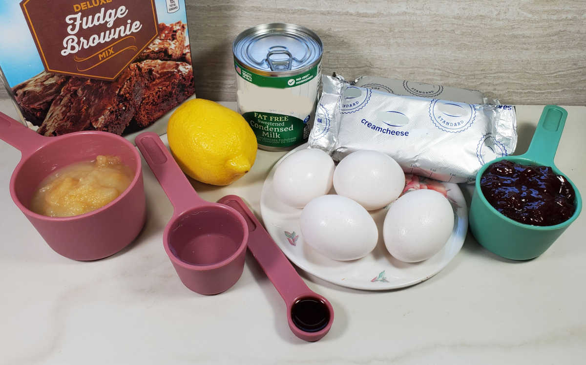 Brownie mix, eggs, lemon, milk, applesauce, cream cheese, jam, extract and water on a counter.