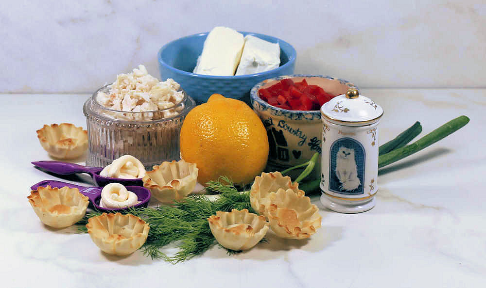 Cream cheese, lemon, red pepper, paprika, horseradish sauce, phyllo cups, dill, onion and crab meat on a counter.