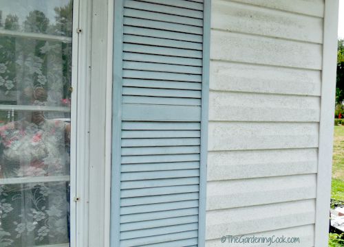 White siding, window with lace curtain and blue shutter.