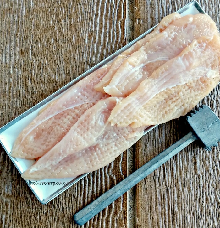 Flattened chicken tenders on a white plate with a meat tenderizer.