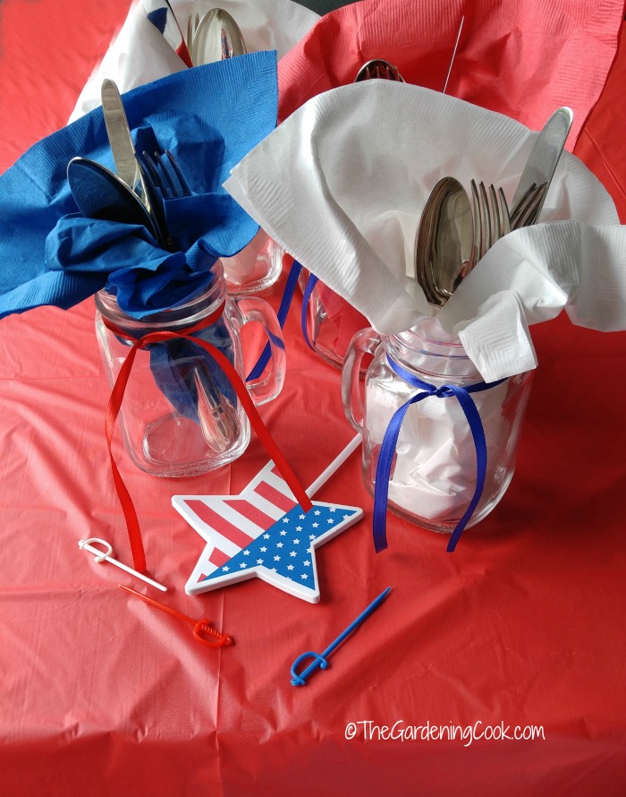 Patiotic Mason Jar cutlery holders with blue and white napkins, red and blue ribbons and flag stars on a red table cloth.