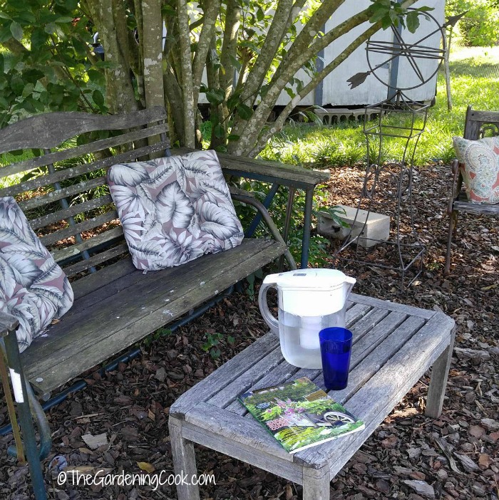 Take plenty of breaks Garden sett8ing with cushions, water jug, glass and magazine on a wooden table.