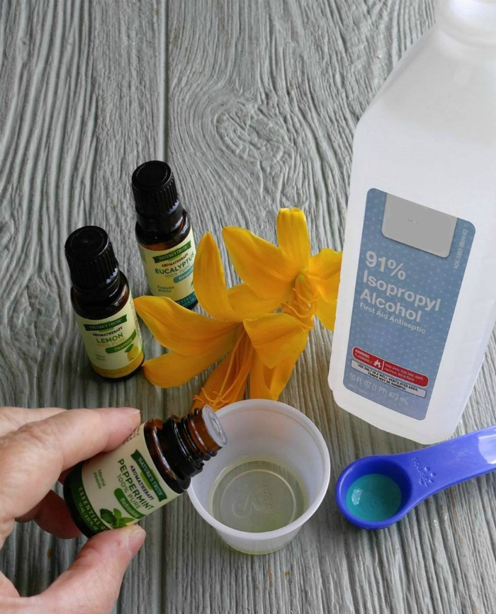 Bottle of rubbing alcohol, flower, teaspoon, three bottles of essential oils and a handing pouring into a measuring cup Add the essential oils to rubbing alcohol