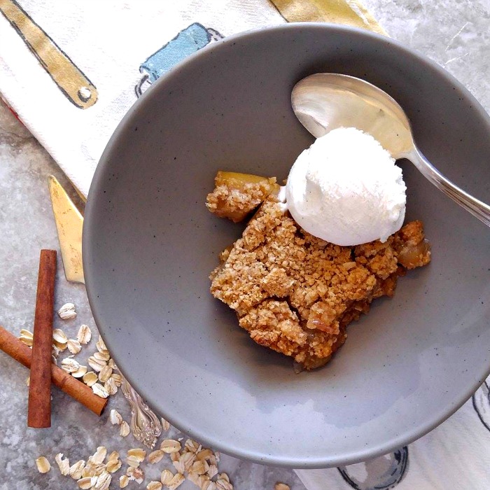 Warm apple pie bars and ice cream in a gray bowl with a spoon, towel, oats and cinnamon