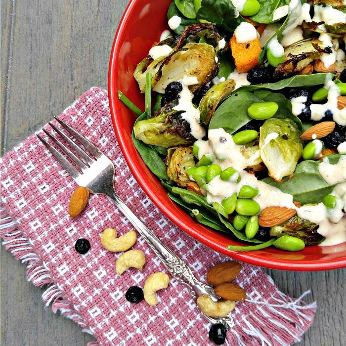 Hearty roast vegetable salad in a red bowl with a fork, pink napkin, blueberries and nuts.
