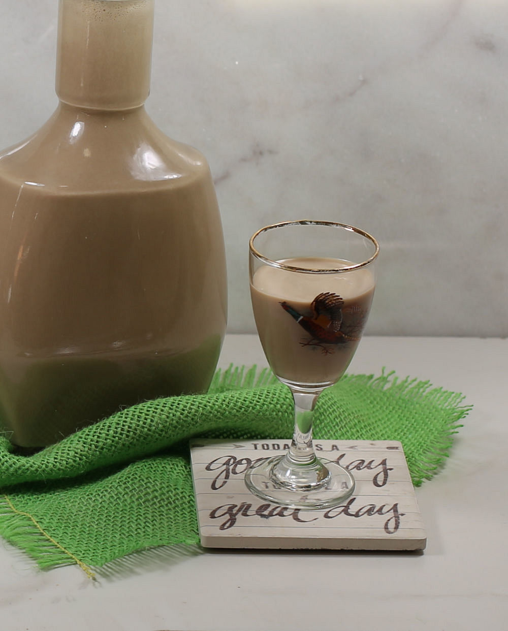 Glass of Baileys Irish cream on a coaster with a green piece of fabric and bottle of Baileys.