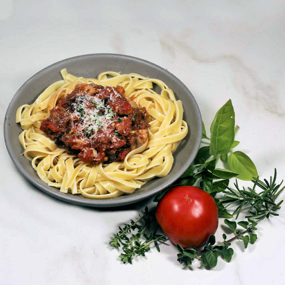 Thick spaghetti sauce with pork and beef near some herbs and a fresh tomato.