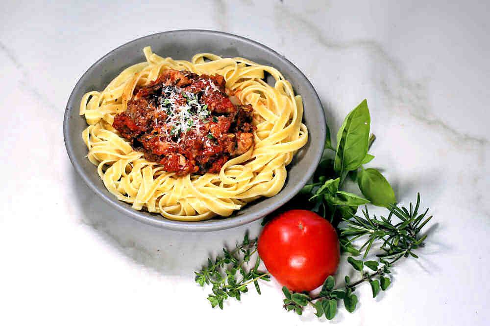 Thick spaghetti sauce with pork and beef over a bowl with fettuccini near some herbs and tomato.